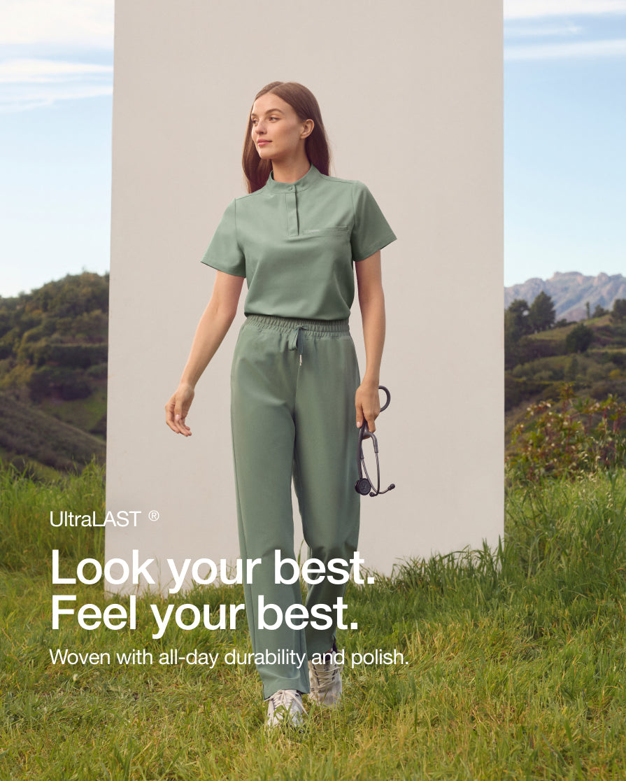 A woman in a sage green Jaanuu UltraLAST scrub set holds a stethoscope on a grassy hillside. Text on the image reads: "Look your best. Feel your best. Woven with all-day durability and polish." with an UltraLAST fabric label.