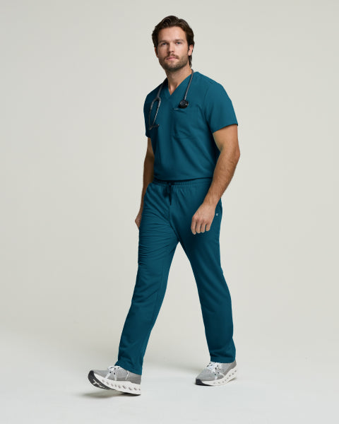 A man in a Caribbean blue Jaanuu scrub set with a stethoscope, against a light gray studio background. - Men Nav Tile