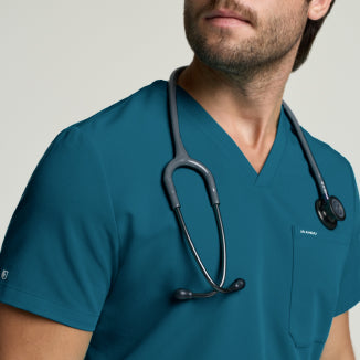 A man in a Caribbean blue Jaanuu scrub set with a stethoscope, against a light gray studio background. - Men Nav Tile