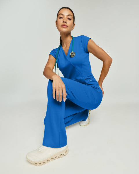 A woman crouching in ceil blue Jaanuu scrubs with a green stethoscope, against a light gray studio background. - Women Nav Tile