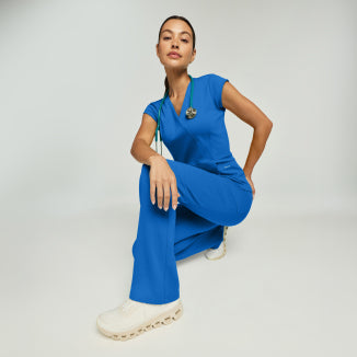 A woman crouching in ceil blue Jaanuu scrubs with a green stethoscope, against a light gray studio background. - Women Nav Tile