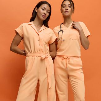 Two women wearing Jaanuu Apricot (peachy orange) scrubs, one in a jumpsuit and one in a scrub top with a stethoscope, on an orange background. - Women Nav Tile