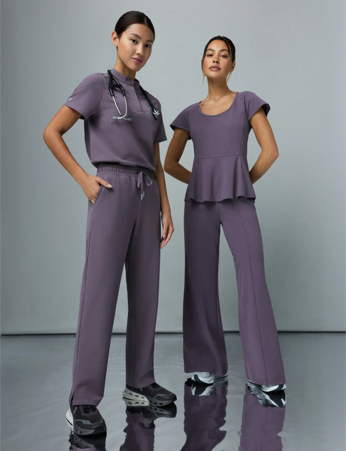 Two women wearing gray titanium Jaanuu scrubs, one in a mock-neck top with a stethoscope, the other in a peplum-style top.