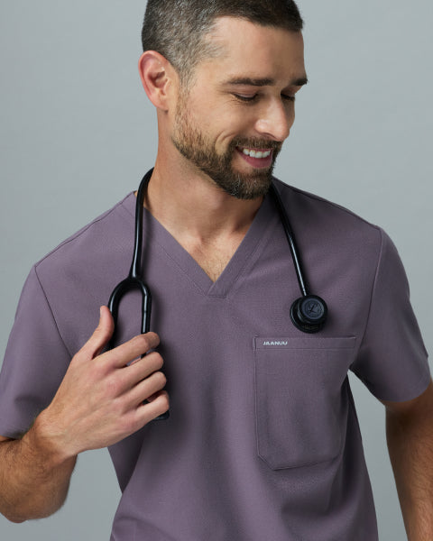 Smiling man in a warm gray Jaanuu Titanium V-neck scrub top holding the stethoscope around his neck, on a light gray background. - Men Nav Tile