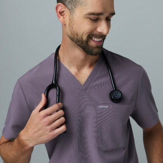 Smiling man in a warm gray Jaanuu Titanium V-neck scrub top holding the stethoscope around his neck, on a light gray background. - Men Nav Tile