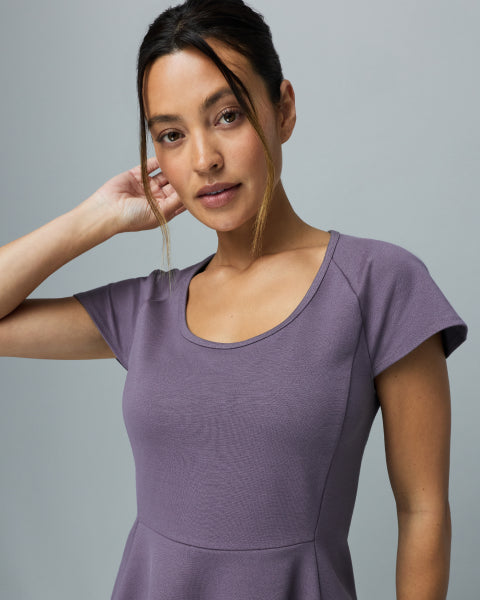 Woman wearing a warm gray Jaanuu Titanium scoop-neck scrub top, hand raised to her hair, on a light gray background. - Women Nav Tile