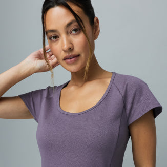 Woman wearing a warm gray Jaanuu Titanium scoop-neck scrub top, hand raised to her hair, on a light gray background. - Women Nav Tile