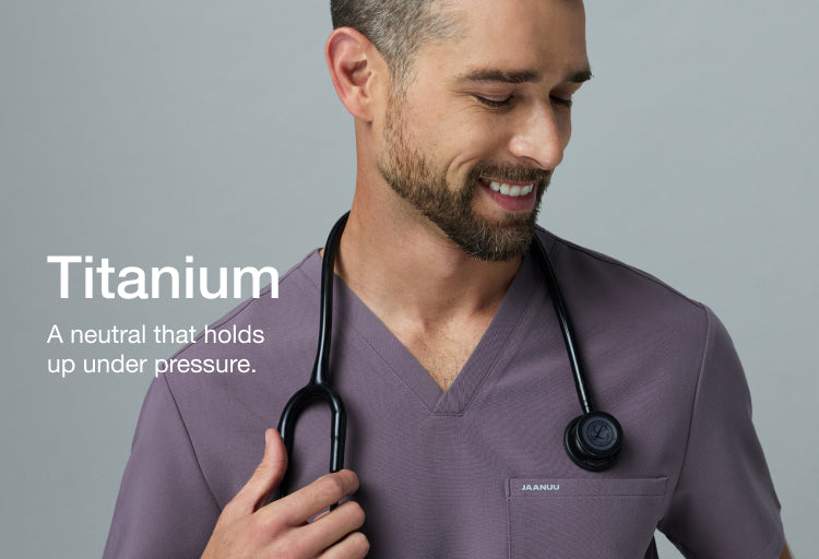 Smiling man in a warm gray Jaanuu Titanium V-neck scrub top with a stethoscope around his neck. Text on the image reads: "Titanium. A neutral that holds up under pressure."