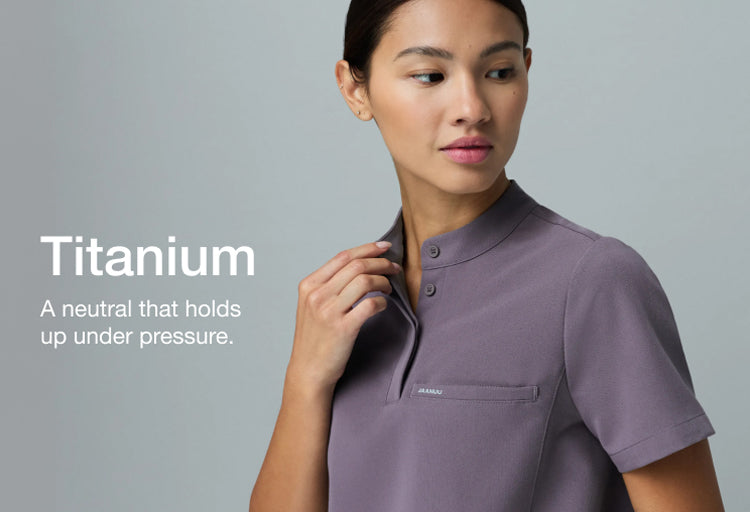 Close-up of a woman in a warm gray Jaanuu Titanium mandarin-collar scrub top, adjusting her collar. Text on the image reads: "Titanium. A neutral that holds up under pressure."