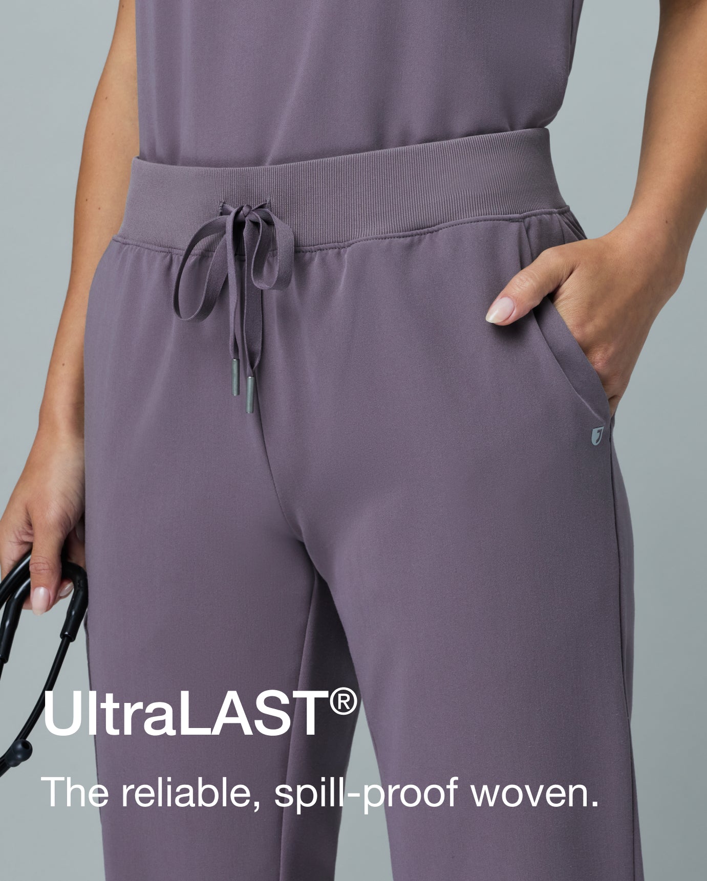 Close-up of a woman in a warm gray Jaanuu Titanium UltraLAST scrub set, hand in her jogger pocket, holding a stethoscope. Text on the image reads: "UltraLAST®. The reliable, spill-proof woven."