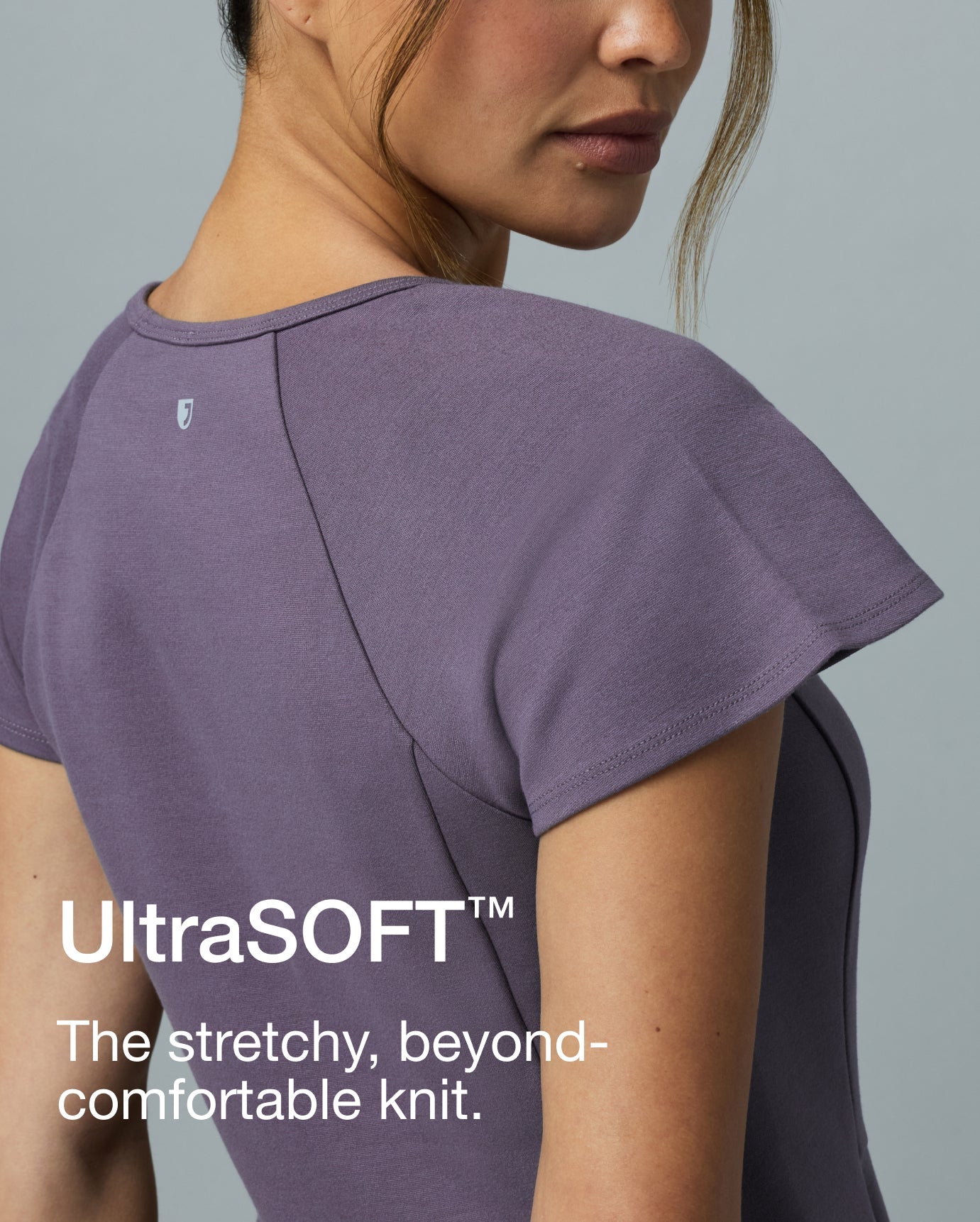 Over-the-shoulder close-up of a woman in a warm gray Jaanuu Titanium UltraSOFT scrub top. Text on the image reads: "UltraSOFT™. The stretchy, beyond-comfortable knit."