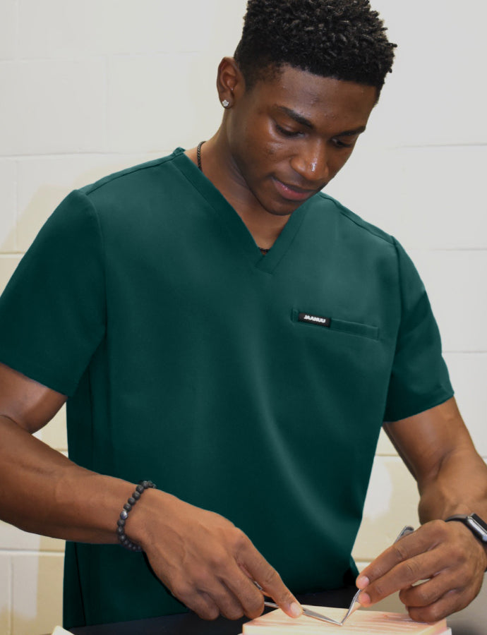 A man in a deep forest green Jaanuu V-neck scrub top with a logo chest pocket practices suturing with forceps on a training pad, against a cream cinderblock wall.
