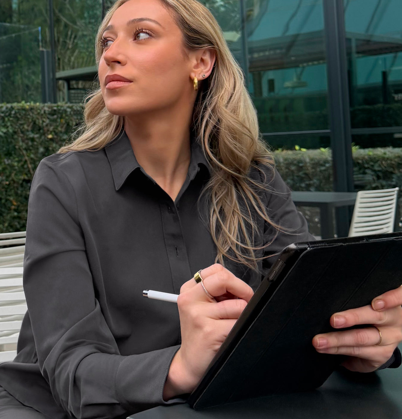 A woman in a dark gray Jaanuu button-front collared top sits outdoors at a table holding a tablet and stylus, looking off to the side, with a glass building and hedge behind her.