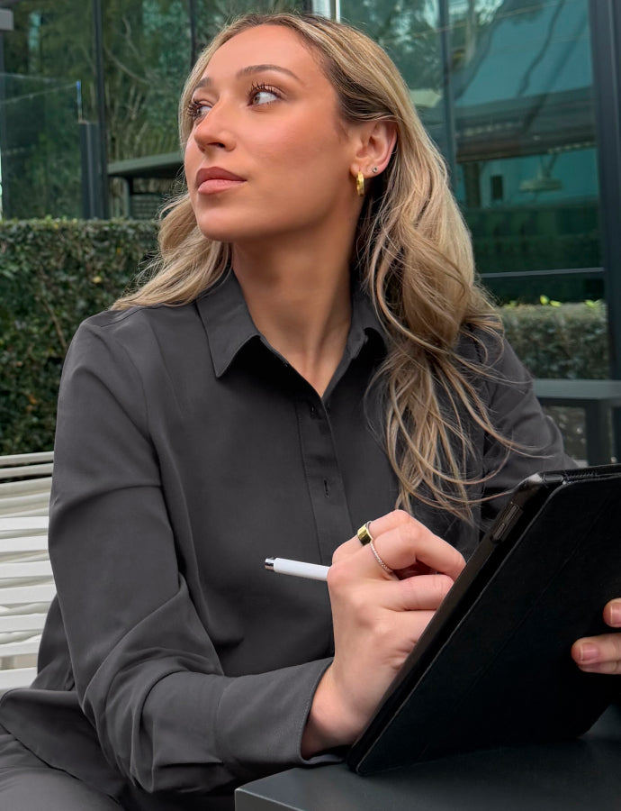 A woman in a dark gray Jaanuu button-front collared top sits outdoors at a table holding a tablet and stylus, looking off to the side, with a glass building and hedge behind her.