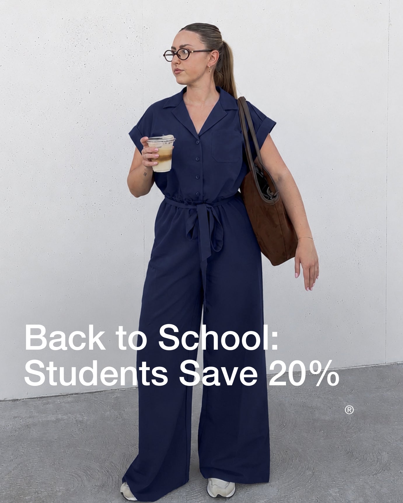 A woman in a Midnight Navy Jaanuu wide-leg scrub jumpsuit with a tie waist stands against a concrete wall, holding an iced coffee with a brown suede tote on her shoulder. Text on the image reads: "Back to School: Students Save 20%"