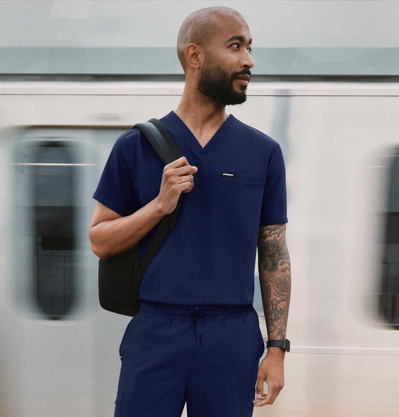 A man wearing scrubs standing in the subway with a train moving in the background.