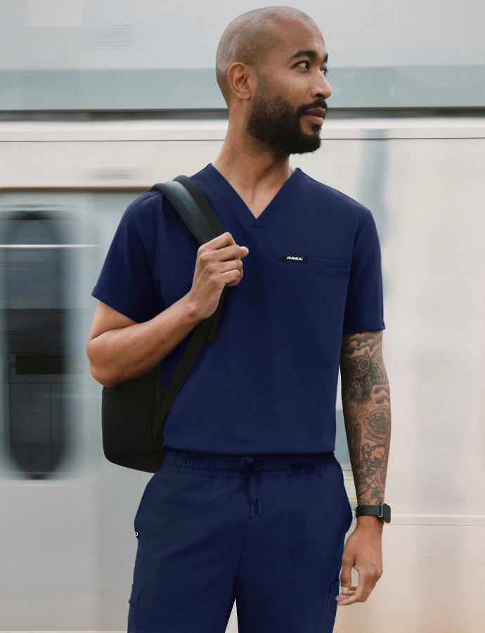 A man wearing scrubs standing in the subway with a train moving in the background.