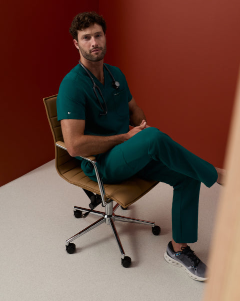 Man in a Midnight Green Jaanuu scrub set with a stethoscope around his neck, sitting in a tan office chair against a rust-red wall - Men Nav Tile