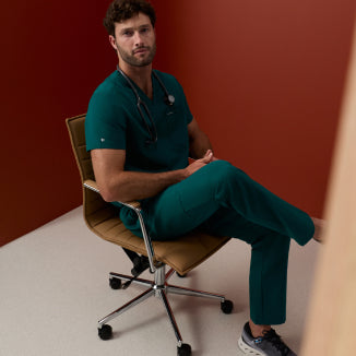 Man in a Midnight Green Jaanuu scrub set with a stethoscope around his neck, sitting in a tan office chair against a rust-red wall - Men Nav Tile