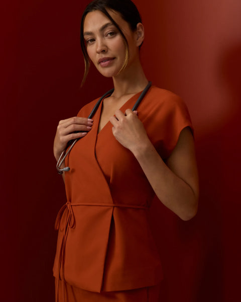 Woman in a Sienna Jaanuu wrap-front scrub top with a stethoscope around her neck, against a deep red background - Women Nav Tile