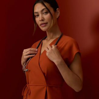 Woman in a Sienna Jaanuu wrap-front scrub top with a stethoscope around her neck, against a deep red background - Women Nav Tile