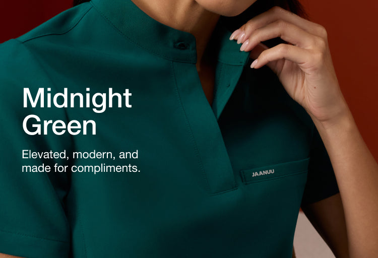 Woman in a Midnight Green Jaanuu scrub top with her hand raised to the collar, against a rust-red background. Text on the image reads: "Midnight Green. Elevated, modern, and made for compliments."