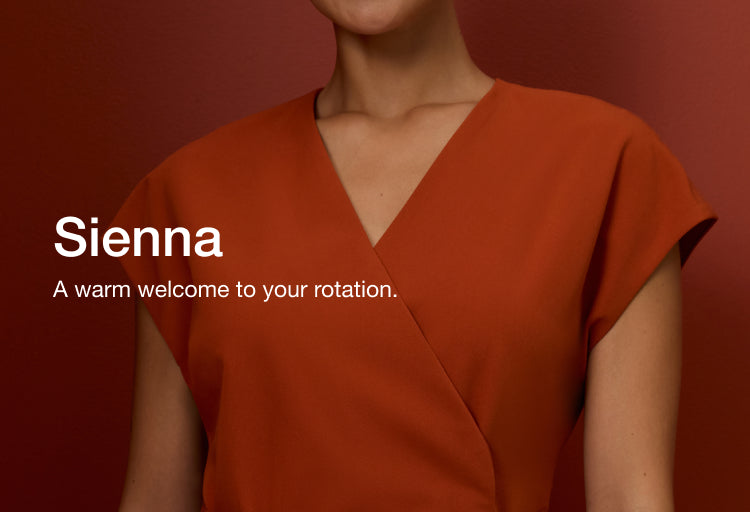 Woman wearing a Sienna Jaanuu wrap-front scrub top against a rust-red wall. Text on the image reads: "Sienna. A warm welcome to your rotation."