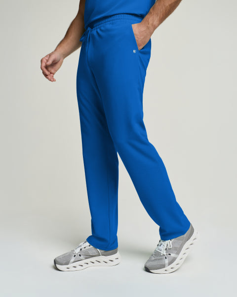 A man walks across a light grey backdrop in royal blue Jaanuu scrub pants and grey sneakers, one hand tucked into the pocket. - Men Nav Tile
