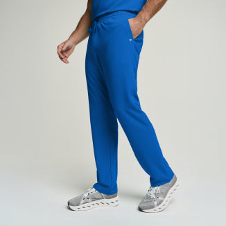 A man walks across a light grey backdrop in royal blue Jaanuu scrub pants and grey sneakers, one hand tucked into the pocket. - Men Nav Tile