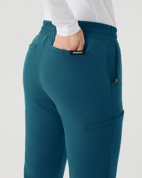 A rear three-quarter view of a woman in teal Jaanuu scrub pants with a white long-sleeve top, one hand tucked into the pocket, against a light grey backdrop. - Women Nav Tile