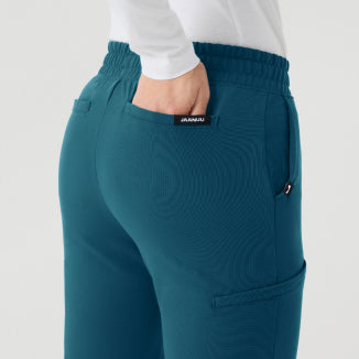 A rear three-quarter view of a woman in teal Jaanuu scrub pants with a white long-sleeve top, one hand tucked into the pocket, against a light grey backdrop. - Women Nav Tile