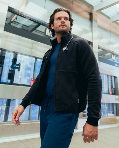 A man in a black Jaanuu fleece zip-front jacket over a navy scrub top and navy scrub pants walks past a glass transit building, a stethoscope around his neck. - Men Nav Tile