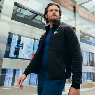A man in a black Jaanuu fleece zip-front jacket over a navy scrub top and navy scrub pants walks past a glass transit building, a stethoscope around his neck. - Men Nav Tile