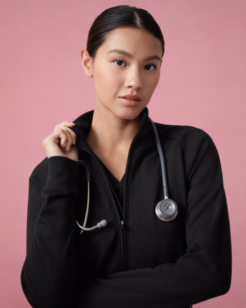 A woman in a black Jaanuu zip-front scrub jacket over a black scrub top stands with her arms crossed and a stethoscope around her neck against a dusty pink backdrop. - Women Nav Tile
