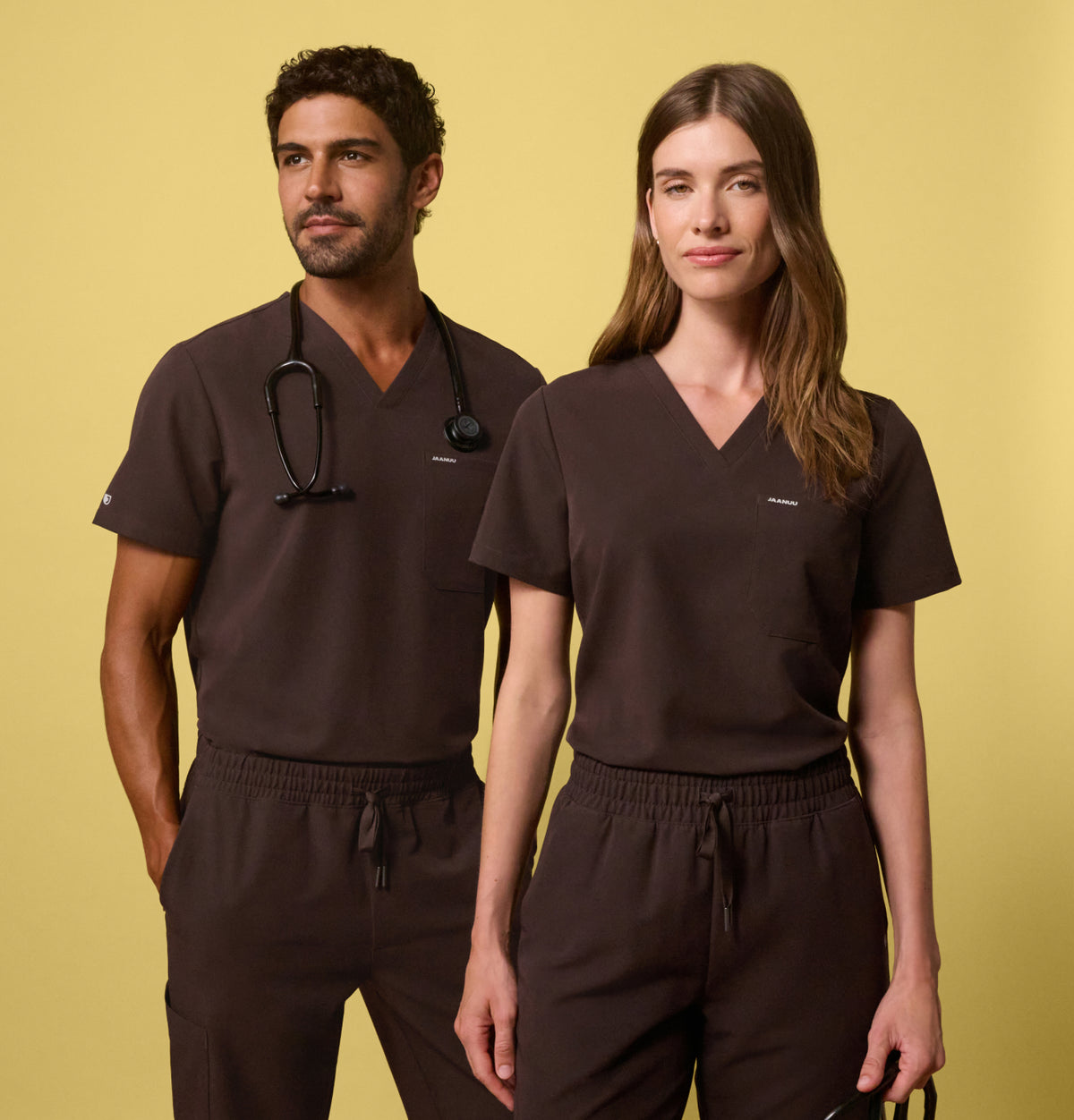A man and woman wearing espresso brown JAANUU scrub sets against a yellow background, one with a stethoscope around the neck and the other holding stethoscope in one hand.