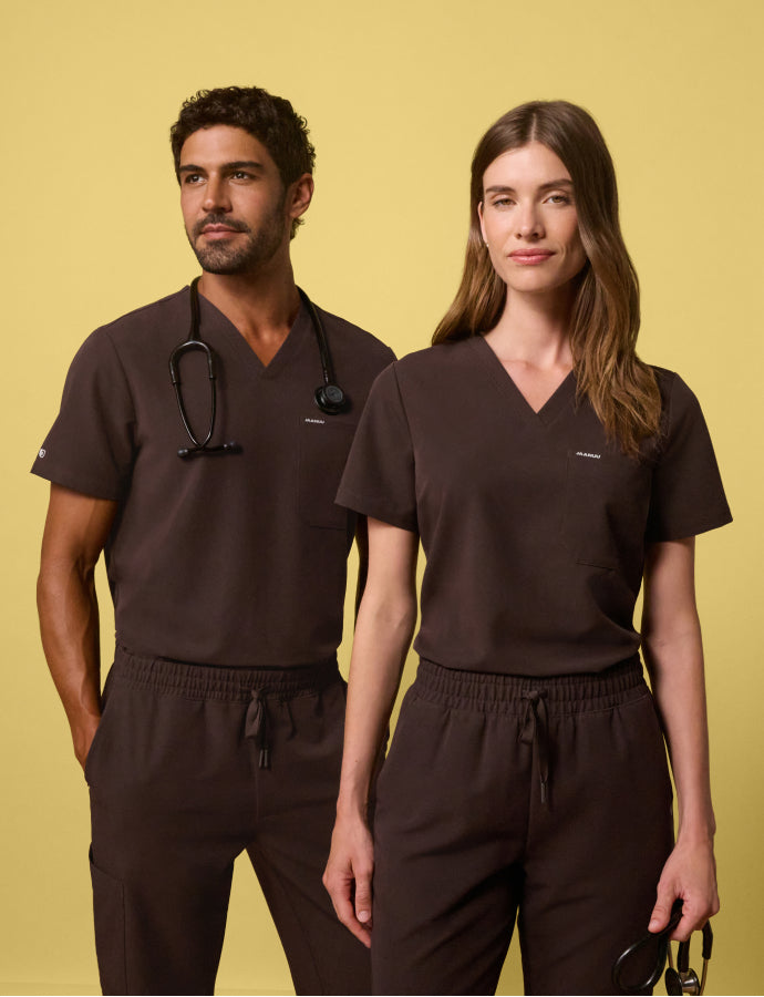 A man and woman wearing espresso brown JAANUU scrub sets against a yellow background, one with a stethoscope around the neck and the other holding stethoscope in one hand.