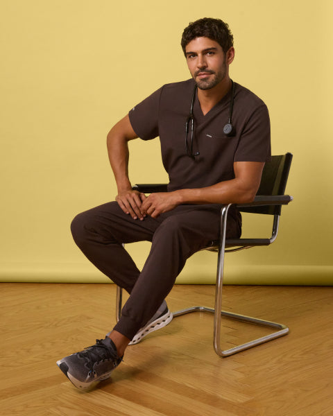 Man seated on a chair wearing Espresso brown scrubs against a yellow background. - Men Nav Tile