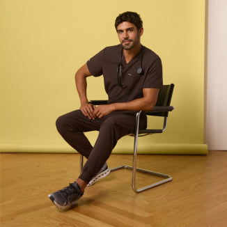 Man seated on a chair wearing Espresso brown scrubs against a yellow background. - Men Nav Tile