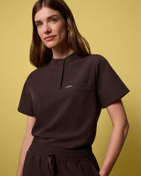 Woman wearing an Espresso brown Savi Stand-Collar scrub top against a yellow background. - Women Nav Tile