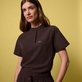 Woman wearing an Espresso brown Savi Stand-Collar scrub top against a yellow background. - Women Nav Tile