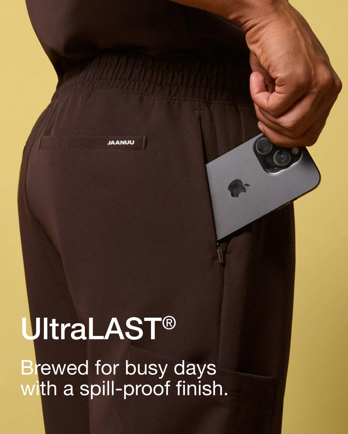 A close-up of a man slipping a phone into the zippered pocket of espresso brown Jaanuu scrub joggers against a yellow backdrop. Text on the image reads: "UltraLAST®. Brewed for busy days with a spill-proof finish."