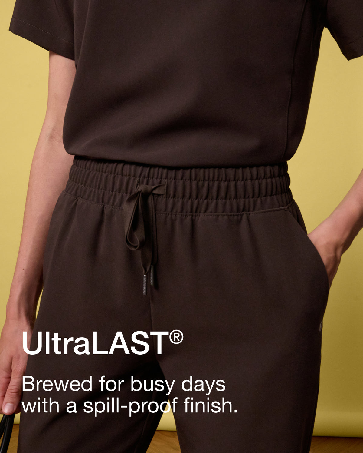 A close-up of a woman in an espresso brown Jaanuu scrub top and drawstring joggers with one hand in the pocket, against a yellow backdrop. Text on the image reads: "UltraLAST®. Brewed for busy days with a spill-proof finish."