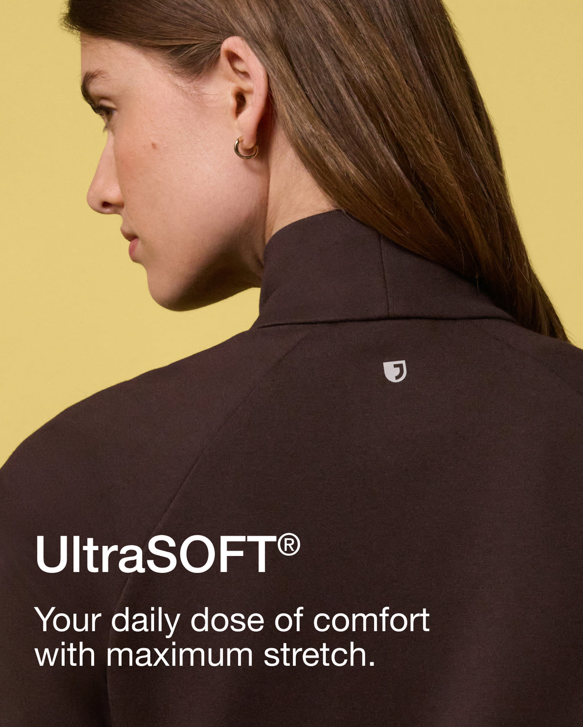 A woman seen from behind in profile wearing an espresso brown Jaanuu mock-neck scrub top against a yellow backdrop. Text on the image reads: "UltraSOFT®. Your daily dose of comfort with maximum stretch."