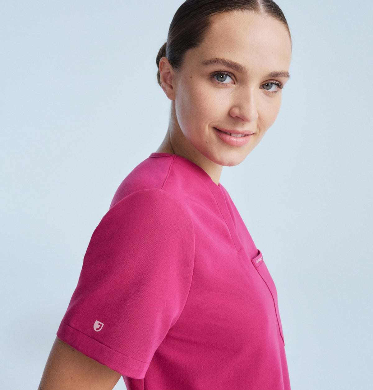 A smiling woman with dark hair pulled back looks over her shoulder at the camera, wearing a Jaanuu Warrior Pink (bright pink) V-neck scrub top with a chest pocket and a small Jaanuu shield logo on the sleeve, against a pale blue-grey background.