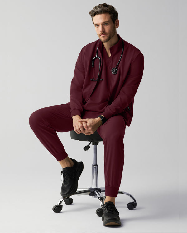 A man in burgundy Jaanuu scrubs sits on a black rolling stool with his hands clasped and a stethoscope around his neck, wearing a zip-up scrub jacket over a V-neck top with matching joggers, against a light grey studio background. - Men Nav Tile