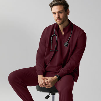 A man in burgundy Jaanuu scrubs sits on a black rolling stool with his hands clasped and a stethoscope around his neck, wearing a zip-up scrub jacket over a V-neck top with matching joggers, against a light grey studio background. - Men Nav Tile