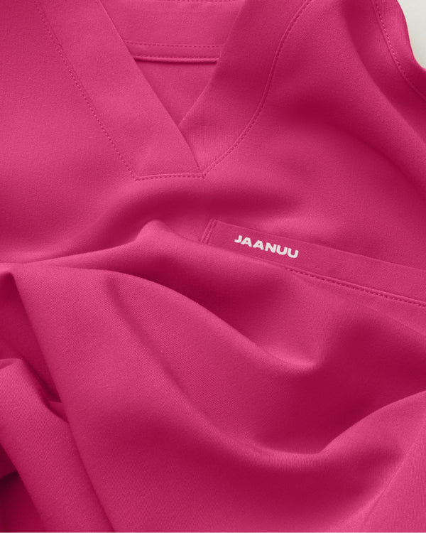 Close-up of a softly folded Jaanuu Warrior Pink (bright pink) V-neck scrub top with a chest pocket and a small white JAANUU logo printed on the pocket. - Women Nav Tile