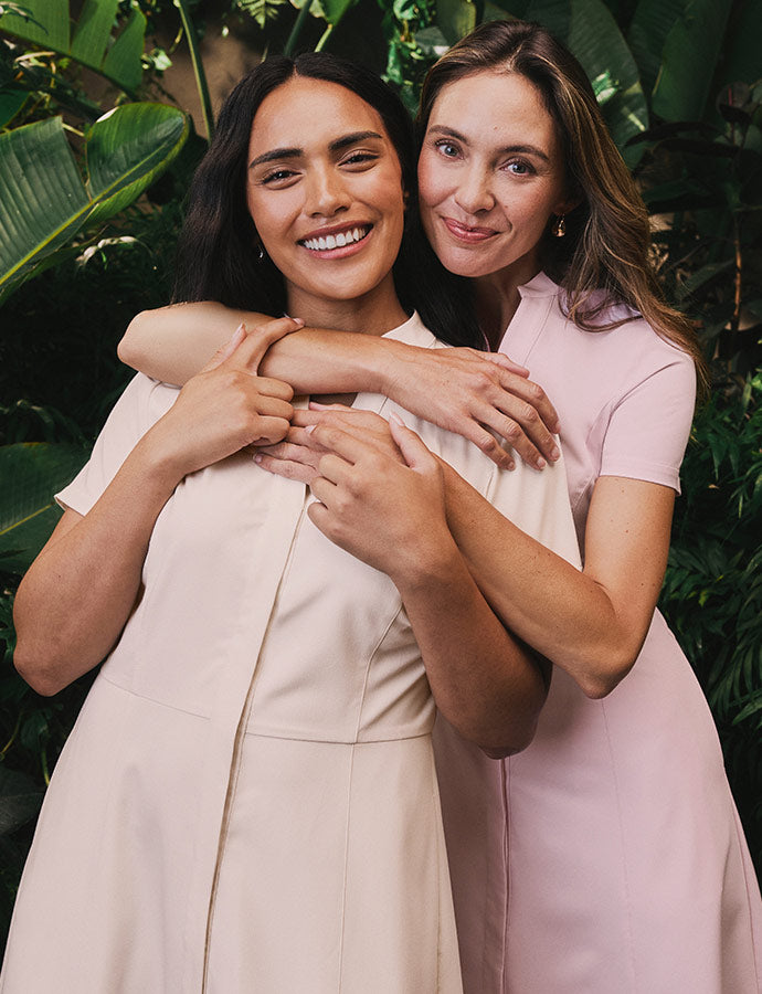 Two women in fashion capsule scrubs, background features tree and plant leaves.