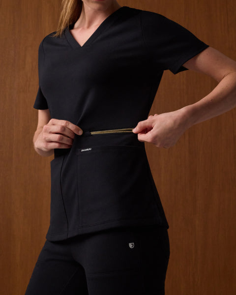 A woman wears a Jaanuu UltraSOFT V-neck scrub top in Black + Gold Zip with matching black scrub pants, against a warm wood-paneled wall, holding the gold zipper that runs across the top's front pocket. - Women Nav Tile