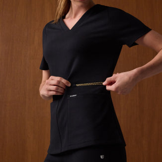 A woman wears a Jaanuu UltraSOFT V-neck scrub top in Black + Gold Zip with matching black scrub pants, against a warm wood-paneled wall, holding the gold zipper that runs across the top's front pocket. - Women Nav Tile